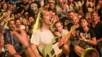 Gitarzysta z długimi włosami grający na elektrycznej gitarze pośród tłumu entuzjastycznych uczestników koncertu, wielu z nich rejestruje wydarzenie telefonami.