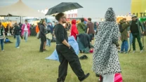 Ludzie na festiwalu ubrani w kolorowe peleryny i płaszcze przeciwdeszczowe, mężczyzna z czarnym parasolem stoi na trawie obok osoby w pelerynie w cętki.