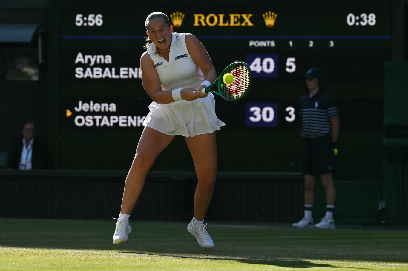 Zawodniczka w białej sukience wykonuje dynamiczny forhend podczas meczu tenisowego na trawiastym korcie, w tle elektroniczna tablica wyników z nazwiskami Aryna Sabalenka i Jelena Ostapenko.