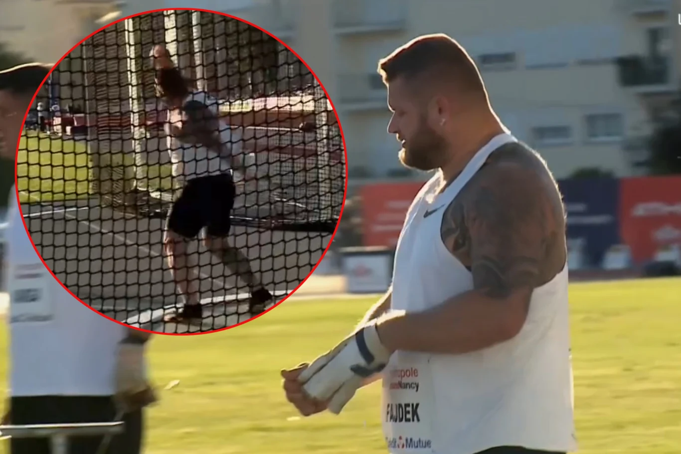 Zawodnik w białej koszulce przygotowuje się do rzutu młotem na stadionie lekkoatletycznym, otoczony przez siatkę ochronną, z widocznym skupieniem na twarzy.