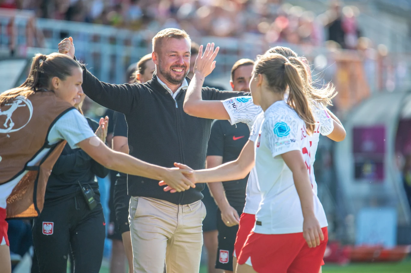 Trener cieszy się razem z piłkarkami reprezentacji Polski po udanym meczu na stadionie, a zawodniczki witają się energicznie z szerokimi uśmiechami.