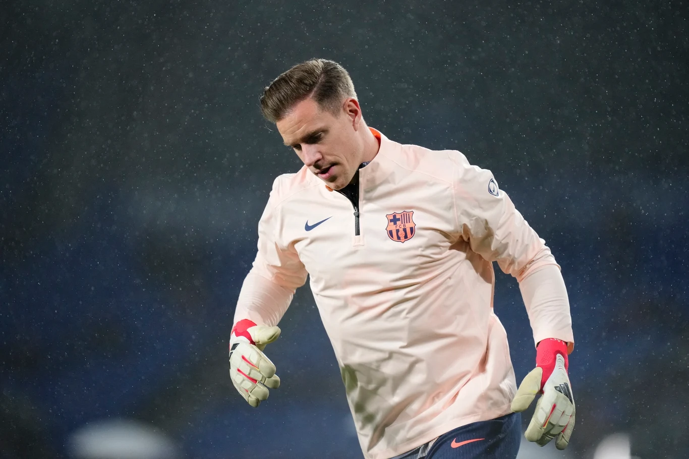 Piłkarz w jasnej bluzie z herbem FC Barcelona oraz rękawicach bramkarskich na tle rozmytego, ciemnego boiska.