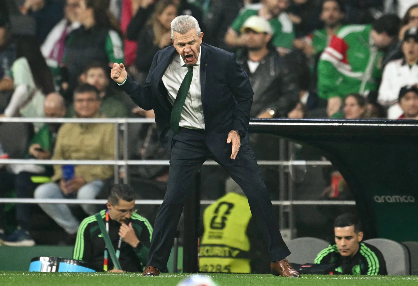 Javier Aguirre Trener w garniturze energicznie gestykuluje przy linii bocznej boiska podczas meczu piłki nożnej, skoncentrowany na wydarzeniach na murawie; obok niego siedzą członkowie sztabu szkoleniowego w zielonych dresach.