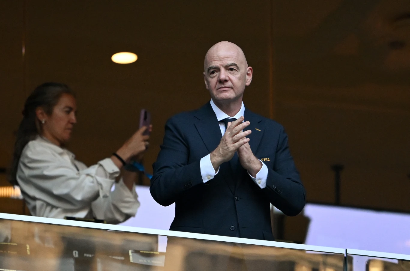 Gianni Infantino Łysy mężczyzna w garniturze stoi w loży, z poważnym wyrazem twarzy splata ręce, obok kobieta robi zdjęcie telefonem.
