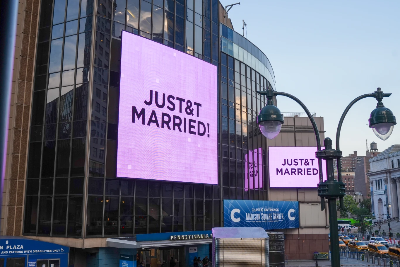 Duży ekran LED z napisem 'JUST&T MARRIED!' zamontowany na elewacji nowoczesnego budynku w centrum miasta, po lewej stronie widoczne wejście do obiektu oraz fragment ruchliwej ulicy z żółtymi taksówkami.