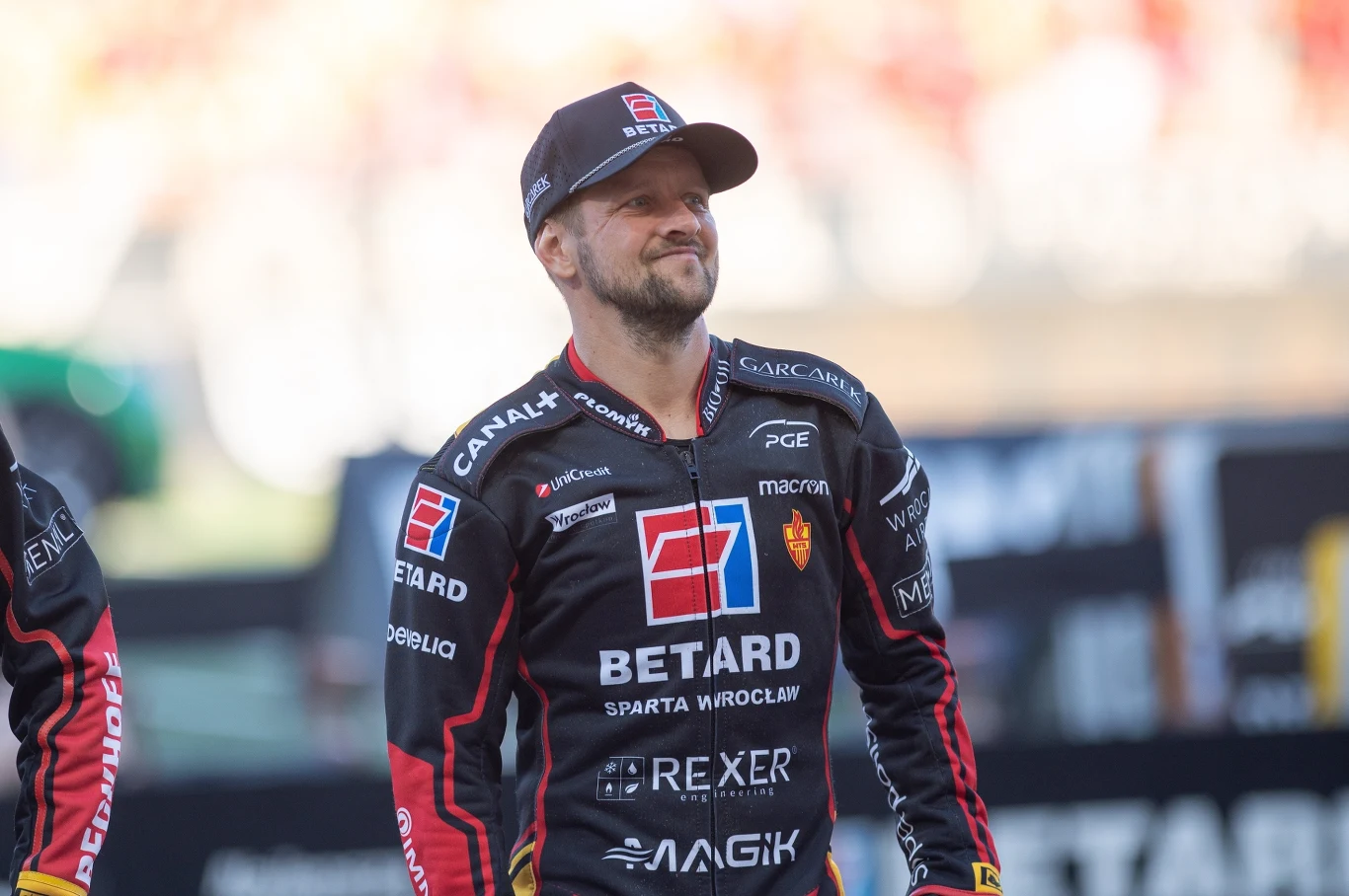 Żużlowiec w czarno-czerwonym kombinezonie zespołu Betard Sparta Wrocław, uśmiechnięty, z daszkiem na głowie, stoi na stadionie sportowym.