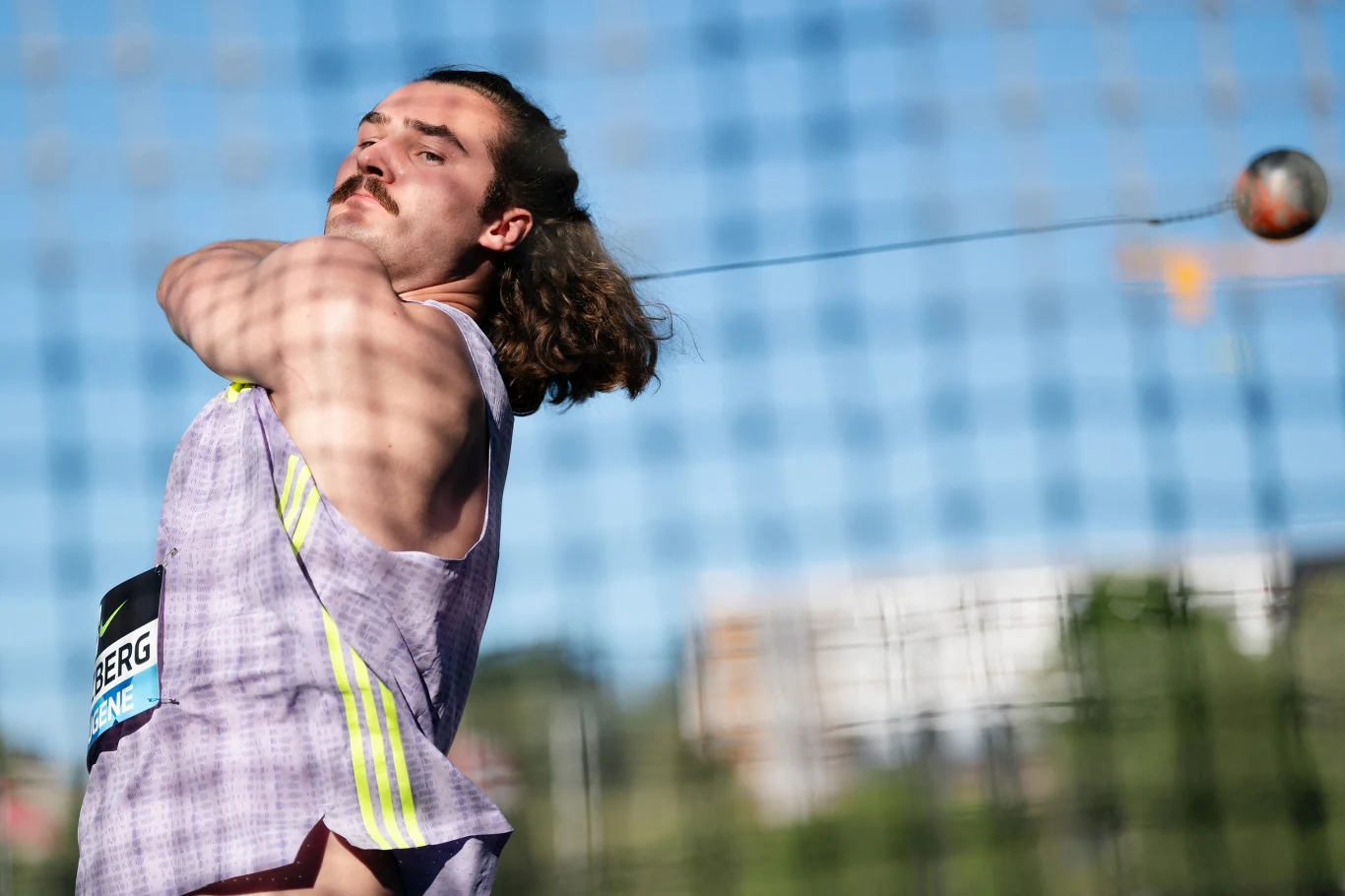 Atleta z długimi włosami wykonuje rzut młotem podczas zawodów lekkoatletycznych, trzymając narzędzie sportowe tuż przed jego uwolnieniem.