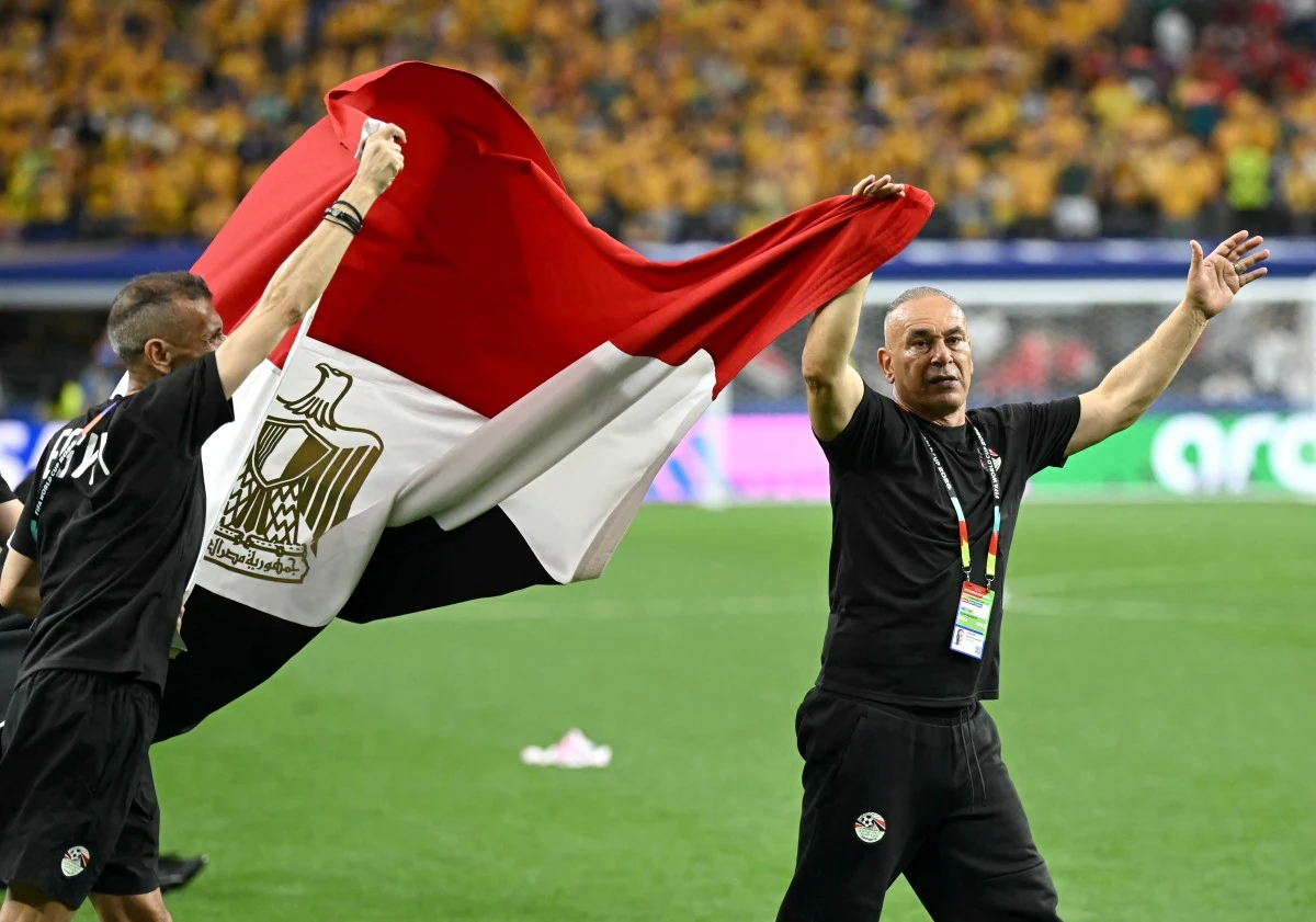 Dwóch mężczyzn ubranych w czarne stroje sportowe unosi dużą flagę Egiptu na stadionie, w tle tłum kibiców ubranych na żółto.
