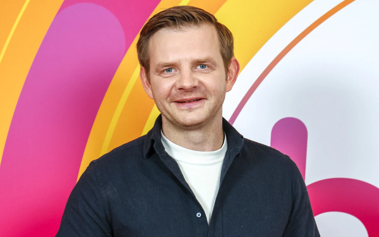 Rafał Zawierucha Uśmiechnięty Rafał Zawierucha w ciemnej koszuli i jasnej koszulce na tle kolorowej ścianki z logo "halo tu polsat"