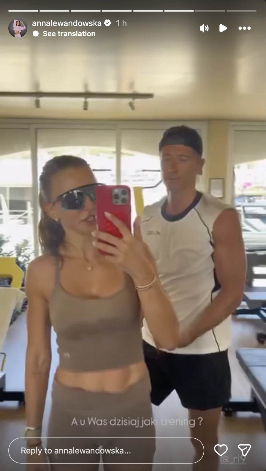 Para ubrana w sportowe stroje podczas treningu na siłowni, kobieta z telefonem robi selfie w dużych okularach przeciwsłonecznych, mężczyzna w białej koszulce i czarnych spodenkach stoi obok.