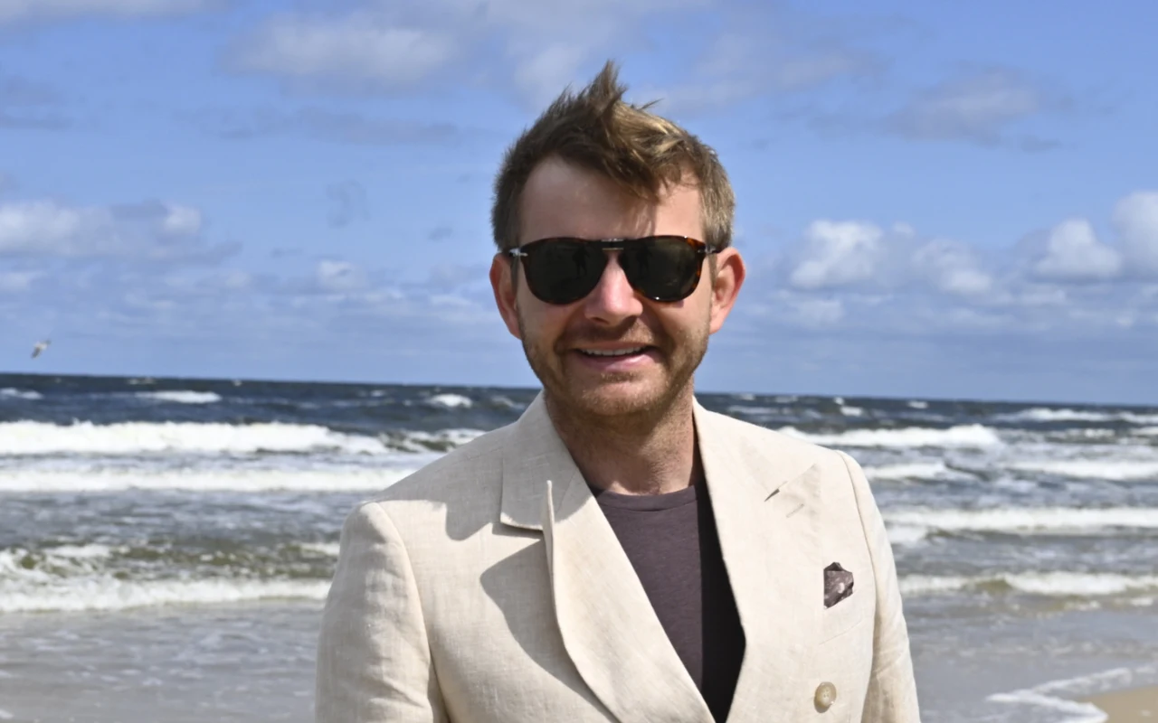 Rafał Zawierucha na 29. Festiwalu Gwiazd w Międzyzdrojach Mężczyzna w jasnej marynarce i okularach przeciwsłonecznych stoi na plaży przy brzegu morza, w tle lekko wzburzona woda i niebo z białymi chmurami.