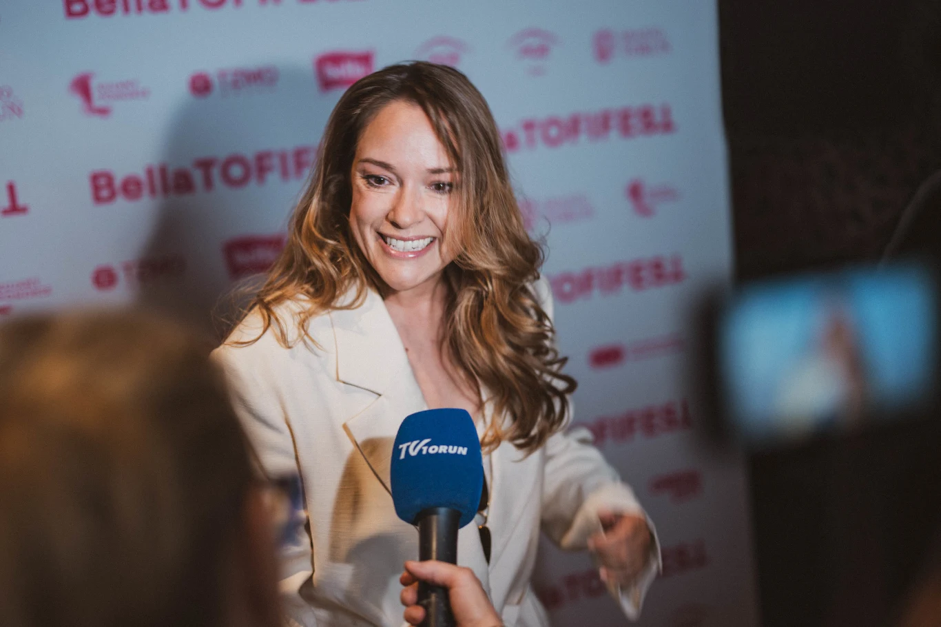 Uśmiechnięta kobieta w jasnej marynarce udziela wywiadu dziennikarzowi trzymającemu niebieski mikrofon TVForum na tle ścianki z logo BellaTOFIFEL.