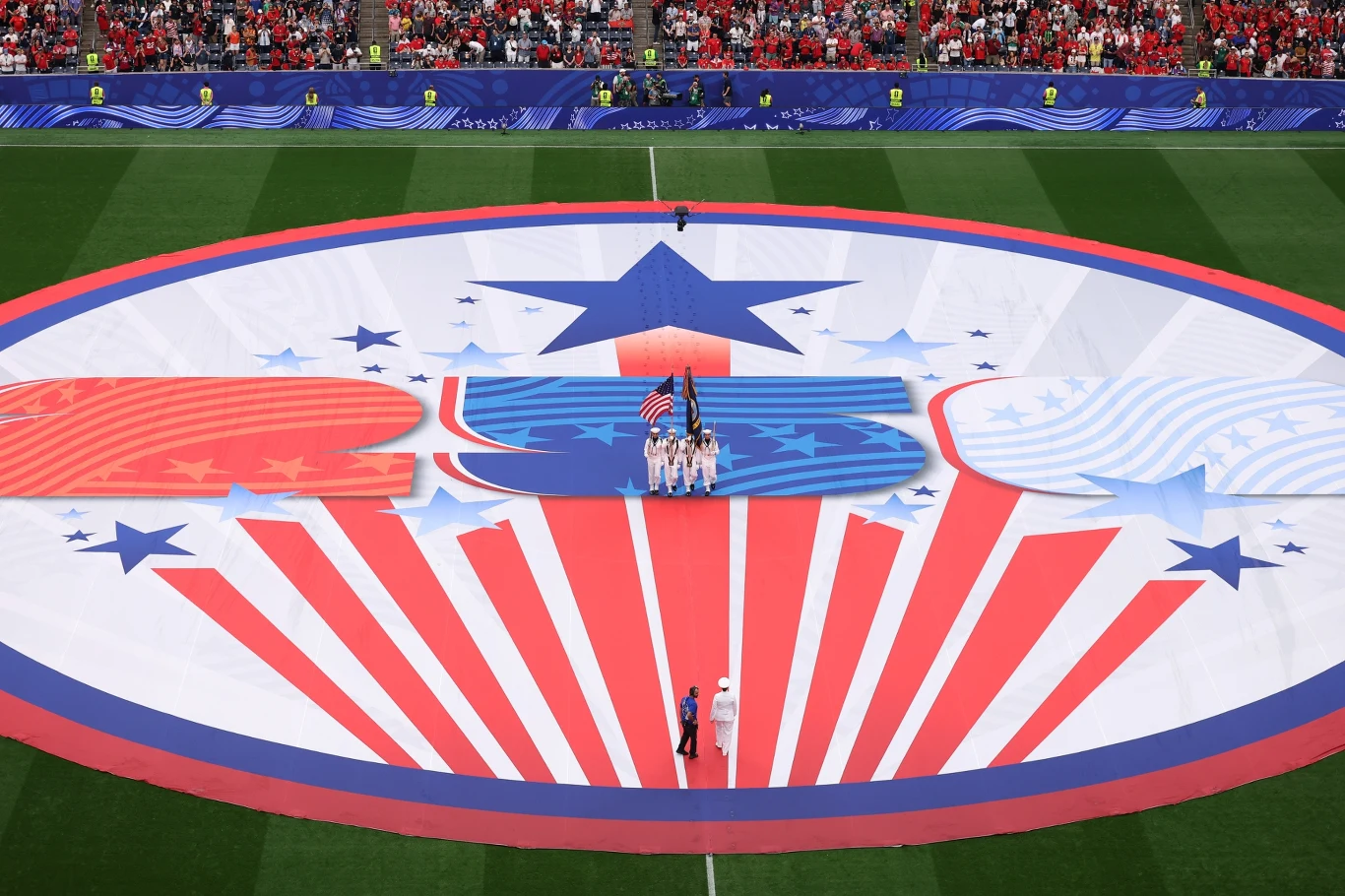 Ogromna, okrągła flaga USA z dominującym motywem gwiazd i pasów leży na murawie stadionu, na jej środku stoi grupa osób trzymających amerykańską flagę narodową, wokół widoczni są kibice na trybunach.