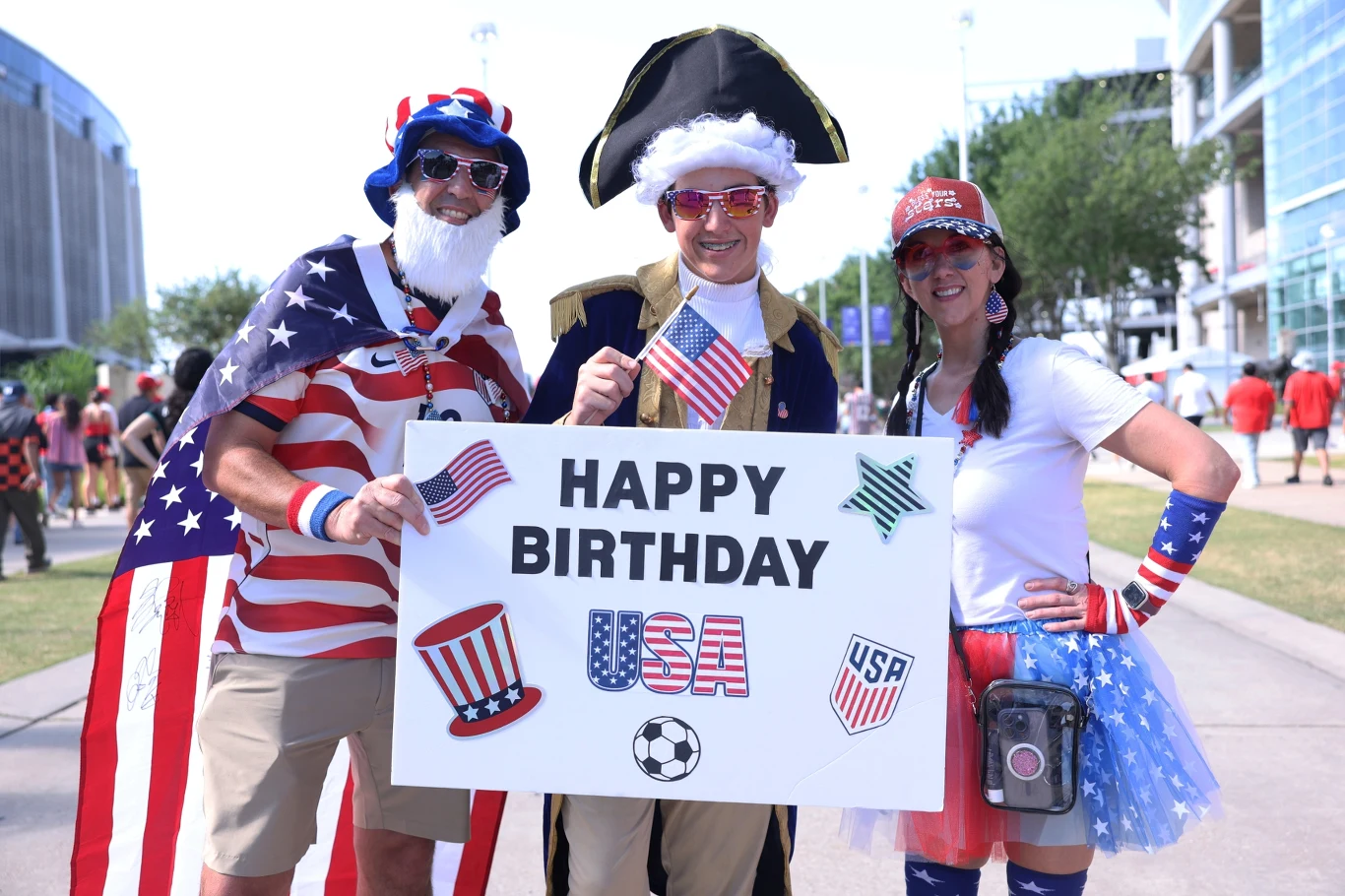Trzy osoby w kolorowych, patriotycznych strojach trzymają ozdobny plakat z napisem „Happy Birthday USA” oraz symbolami Stanów Zjednoczonych, stojąc na tle jasnej miejskiej przestrzeni.