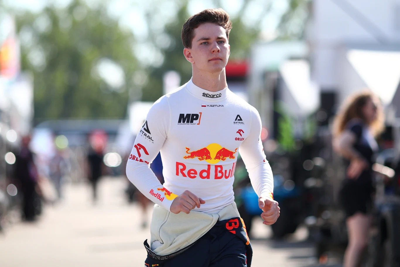 Młody mężczyzna w białym stroju wyścigowym z logotypem Red Bull maszeruje zdecydowanym krokiem po terenie wyścigowym.