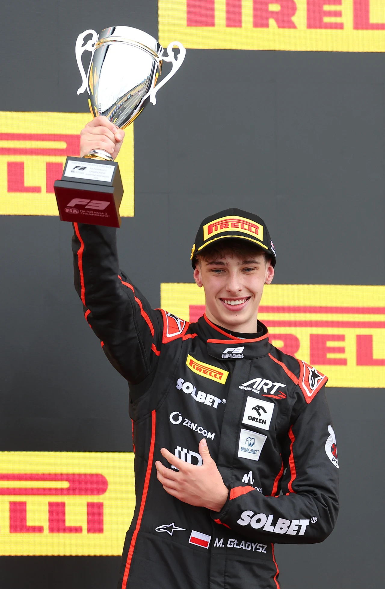 Uśmiechnięty kierowca wyścigowy w czarnym kombinezonie z licznymi sponsorami, trzymający w górze srebrny puchar na podium z tłem ozdobionym logo Pirelli.