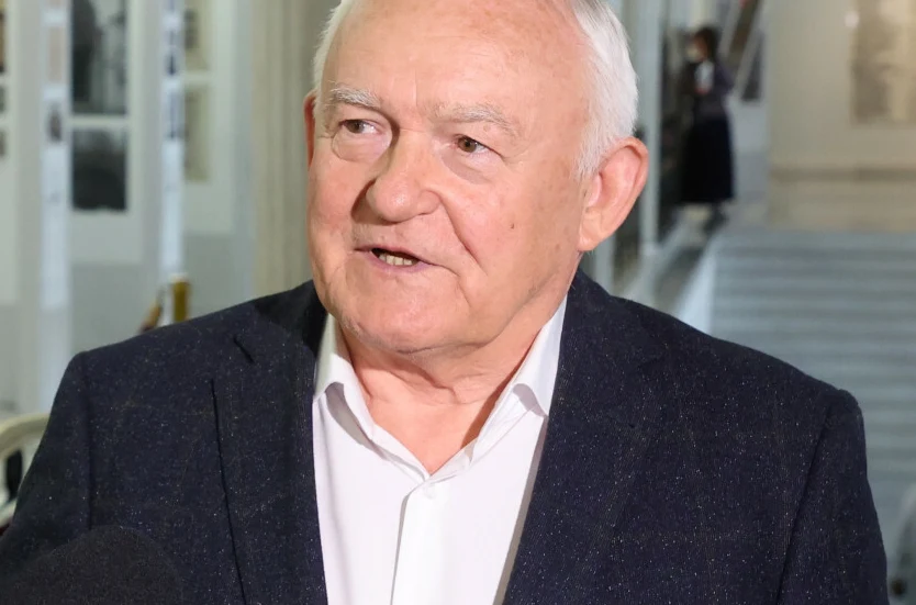 Leszek Miller Leszek Miller w białej koszuli i ciemnej marynarce stoi w jasnym pomieszczeniu.