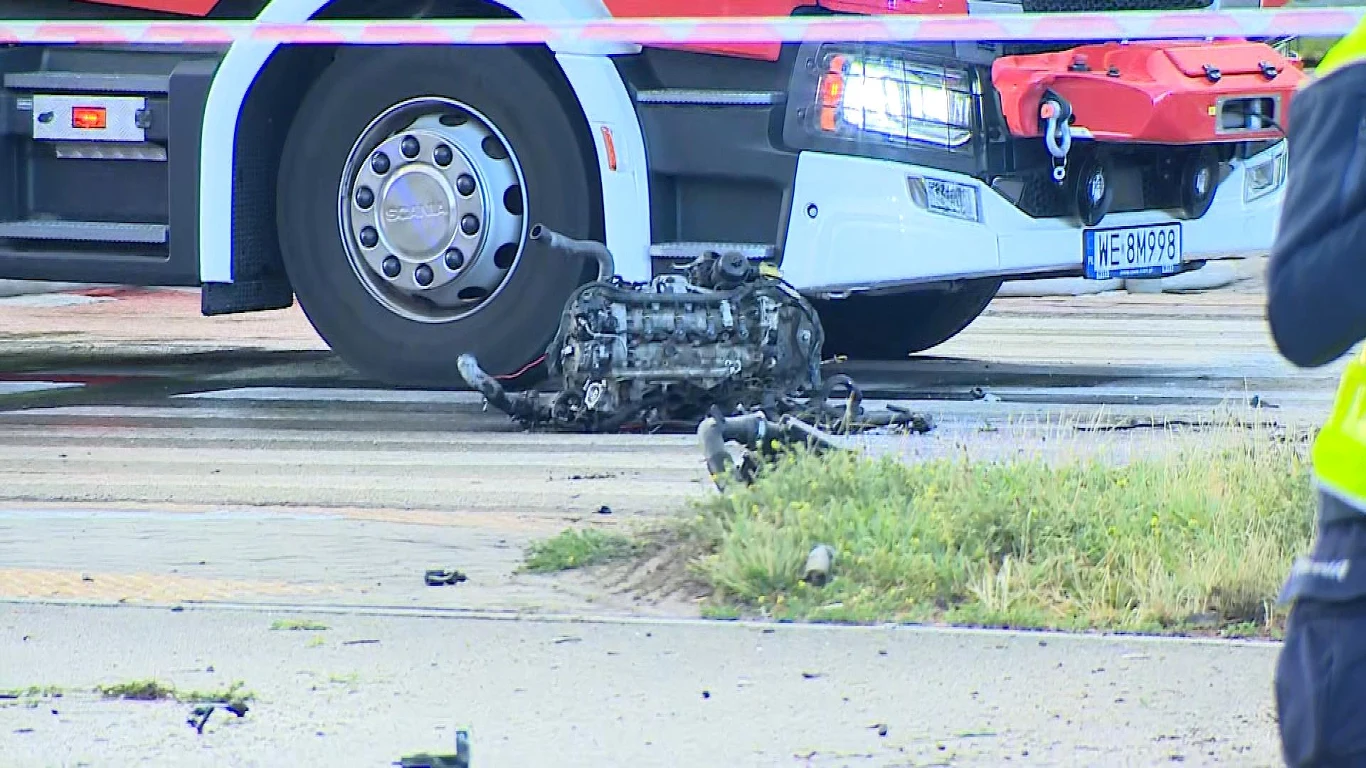 Fragment pojazdu na drodze po wypadku z udziałem autobusu miejskiego Uszkodzony silnik leżący na asfalcie w pobliżu pojazdu służb ratunkowych oraz taśmy zabezpieczającej miejsce zdarzenia.