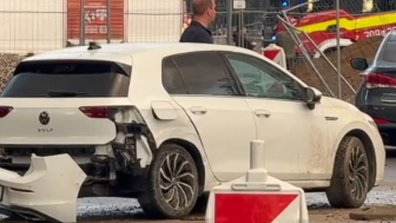 Samochód uszkodzony w wyniku wypadku autobusu miejskiego Biały samochód osobowy z poważnie uszkodzonym tyłem stoi na placu budowy, obok pojazdu znajduje się mężczyzna, a w tle widoczny jest pojazd straży pożarnej.