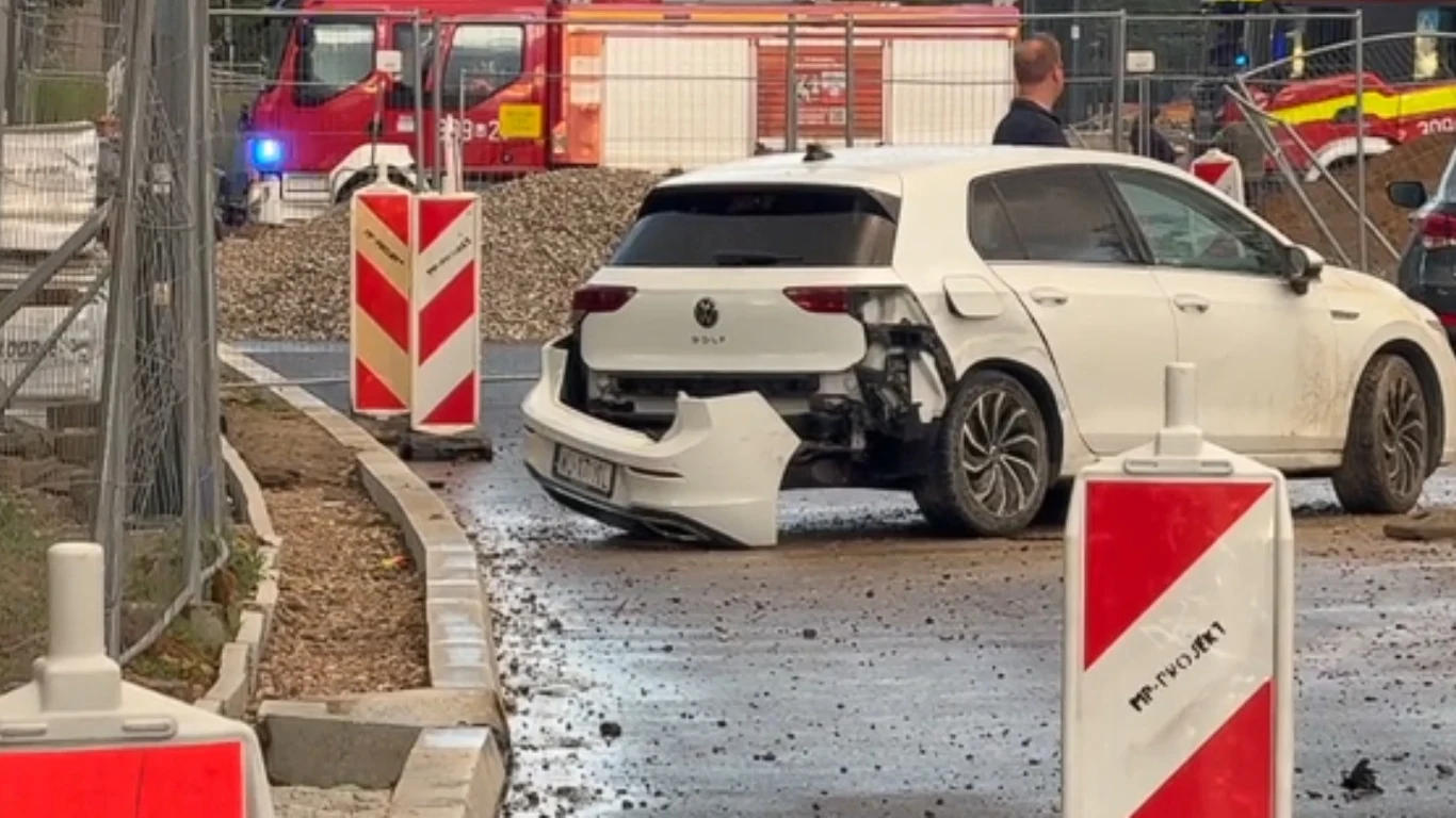Uszkodzony biały samochód osobowy z oderwanym tylnym zderzakiem stoi na drodze wśród ustawionych przy remontowanym odcinku ulicy barier drogowych, w tle pojazdy straży pożarnej i osoba obserwująca sytuację.