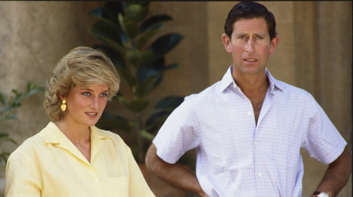 Księżna Diana i ówczesny książę Karol w 1987 roku Dwie elegancko ubrane osoby stoją obok siebie, jedna w żółtej koszuli i żółtych kolczykach, druga w jasnej koszuli z krótkim rękawem, w tle rozmyta roślina o dużych liściach.