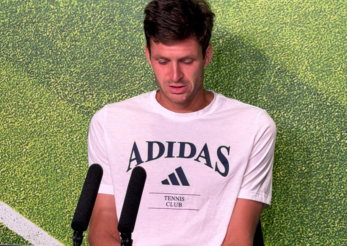 Mężczyzna w białej koszulce Adidas Tennis Club siedzi przed dwoma mikrofonami na tle zielonego boiska tenisowego.