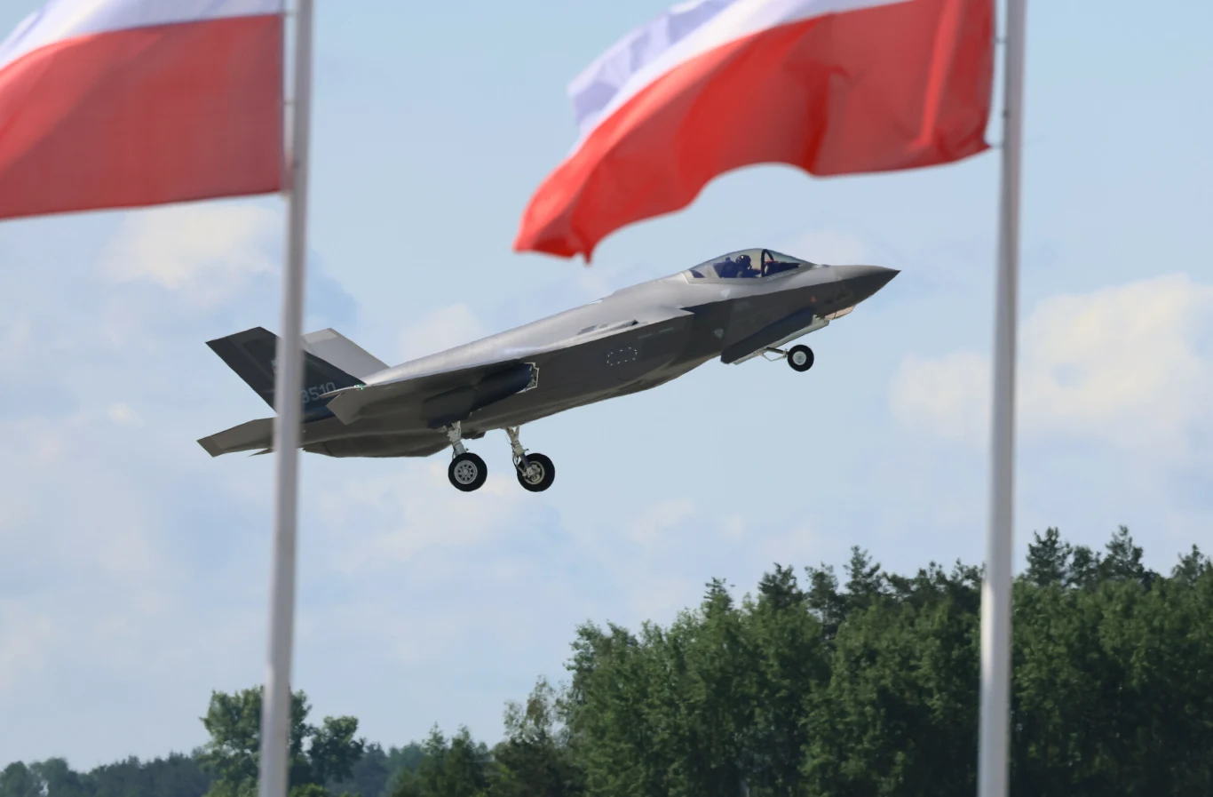 Polskie myśliwce F-35 (zdj. ilustracyjne) Myśliwiec F-35 podczas startu, na pierwszym planie dwie polskie flagi, w tle drzewa i zachmurzone niebo