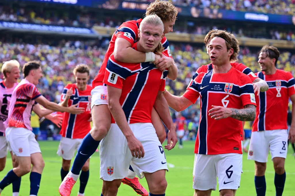 Piłkarze norweskiej reprezentacji w czerwonych strojach świętują sukces na boisku; jeden z zawodników niesie kolegę na plecach, a wokół nich towarzysze z drużyny cieszą się ze zwycięstwa.