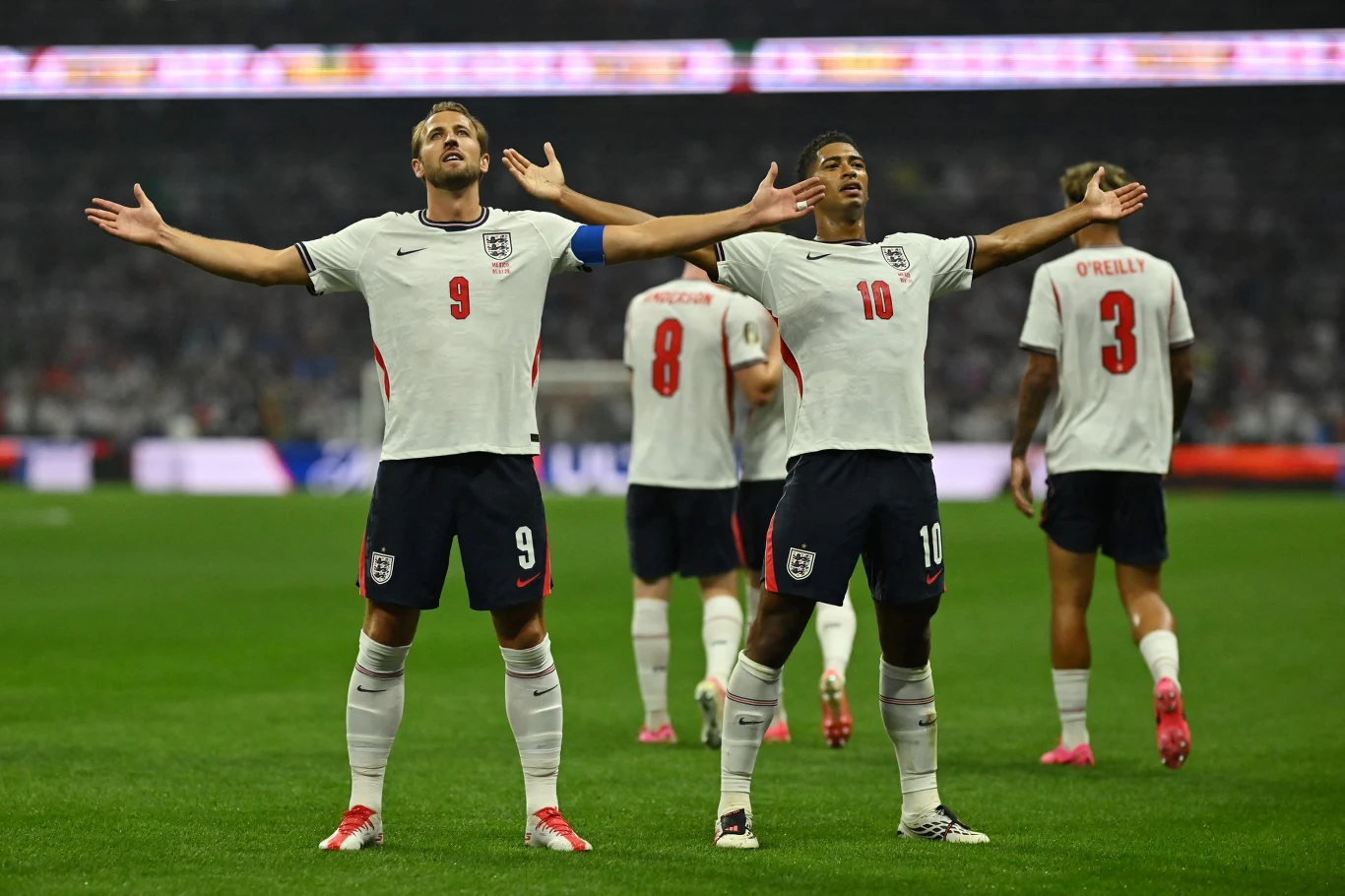 Mundial 2026: Harry Kane i Jude Bellingham Dwóch piłkarzy w białych strojach z numerami 9 i 10 stoi wyprostowanych na murawie stadionu z uniesionymi ramionami, za nimi widać kolejnych członków tej samej drużyny narodowej.