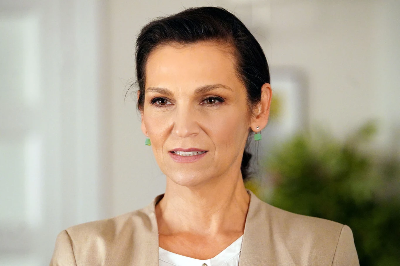 Kobieta o ciemnych włosach upiętych do tyłu w beżowej marynarce, z zielonymi kolczykami i delikatnym makijażem, na neutralnym tle z rozmytymi elementami roślinnymi.