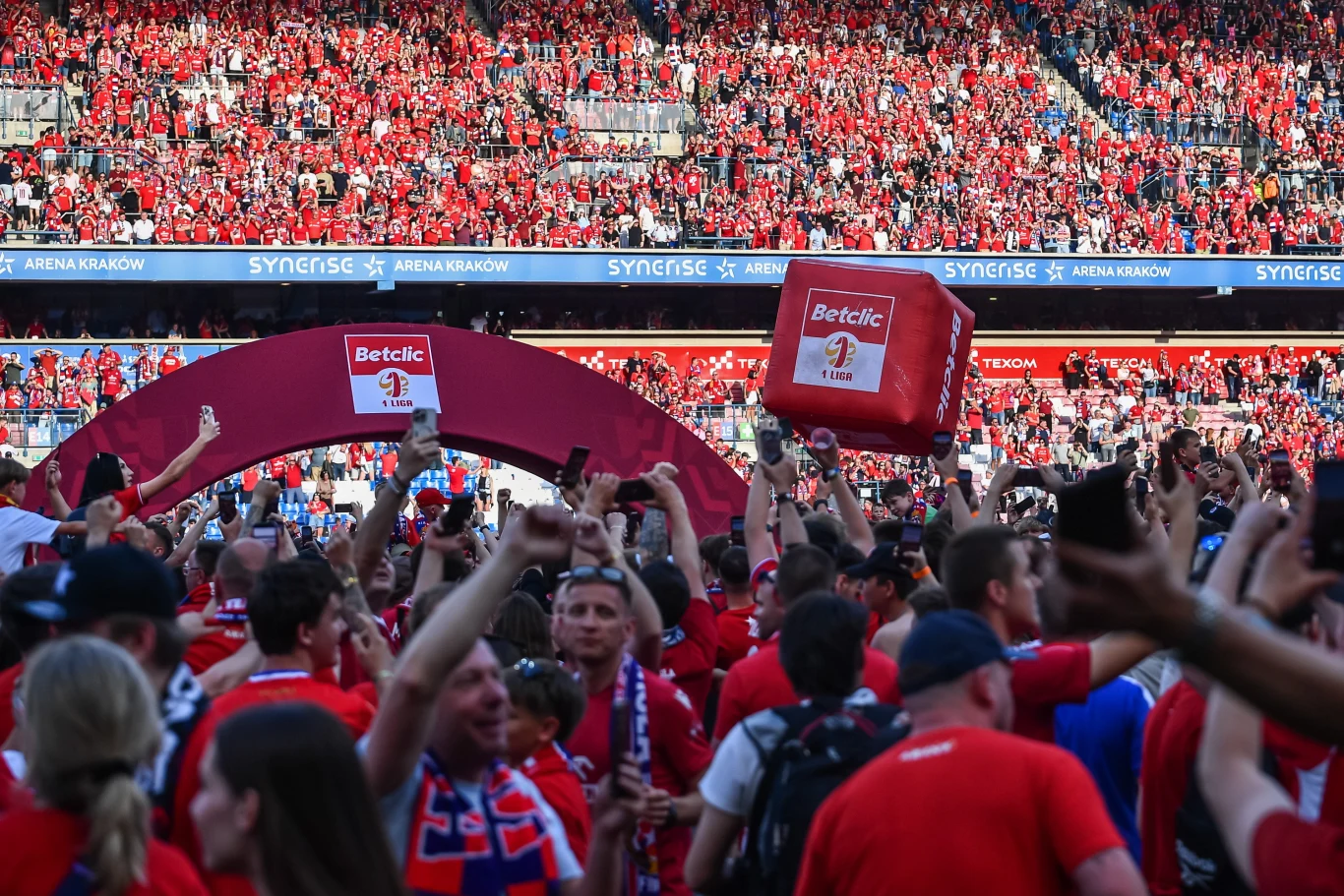 Tak kibice Wisły Kraków świętowali awans do Ekstraklasy Kibice Wisły Kraków ubrani na czerwono świętują awans na stadionie pod łukiem z logo sponsora z dmuchanymi gadżetami.