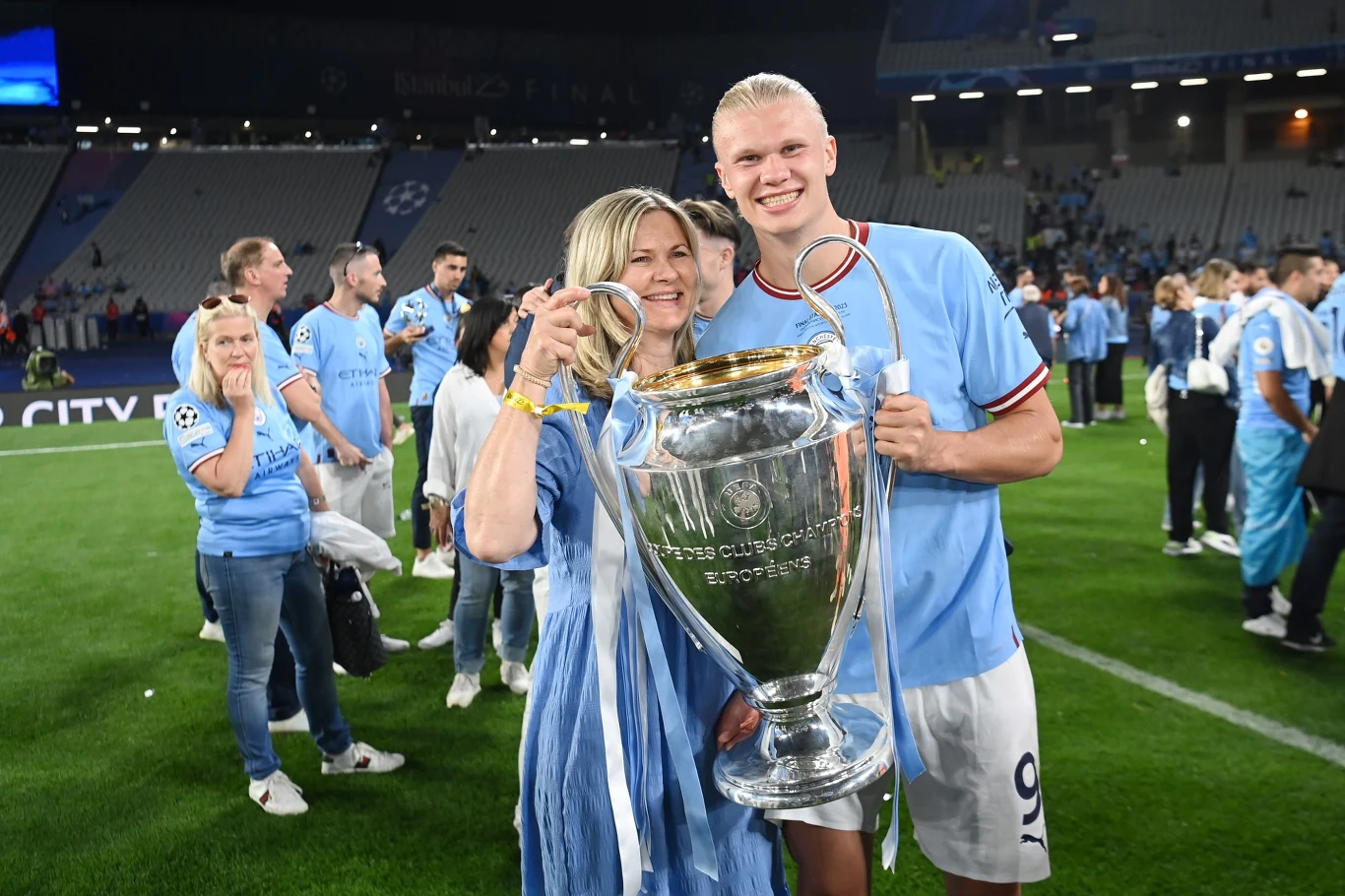 Zawodnik w niebieskiej koszulce stoi z kobietą na boisku, razem trzymają dużą srebrną trofeum piłkarską w otoczeniu innych osób celebrujących zwycięstwo.