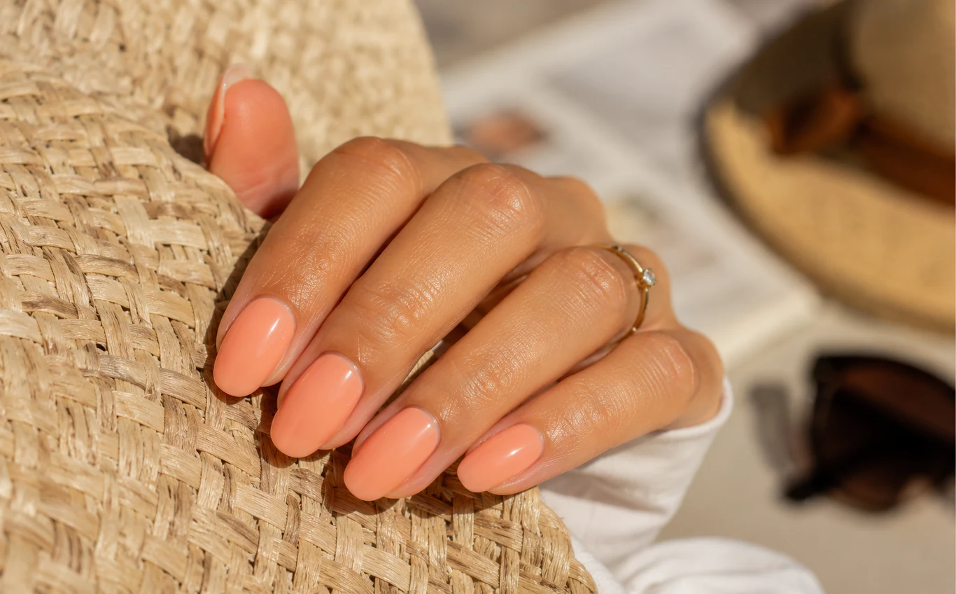 Milky peach to kolor, który pasuje każdemu Dłoń z delikatnym pierścionkiem prezentuje paznokcie pomalowane na pastelowy, brzoskwiniowy kolor, oparta o jasny słomkowy kapelusz.
