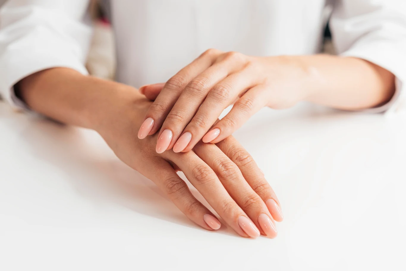 Brzoskwiniowe paznokcie to prawdziwy hot wakacji Zadbane dłonie z naturalnie wyglądającym manicure, ułożone jedna na drugiej na białej powierzchni.