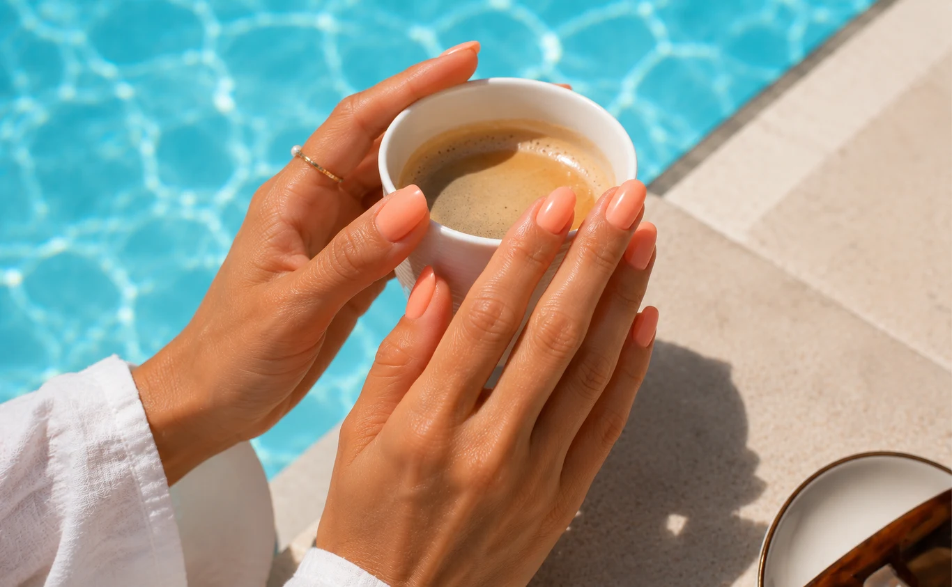 Manicure, który odmładza dłonie Dłonie z naturalnym manicure trzymające filiżankę kawy nad brzegiem basenu z błękitną wodą.
