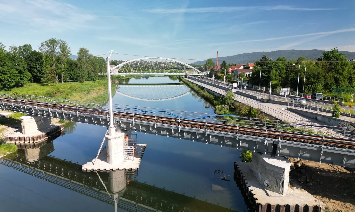 Koleją szybciej w Beskidy. PKP PLK kończy modernizację linii nr 97. Linia kolejowa nr 97 przebiegająca przez rzekę, most w trakcie budowy, zieleń i zabudowania po prawej stronie.
