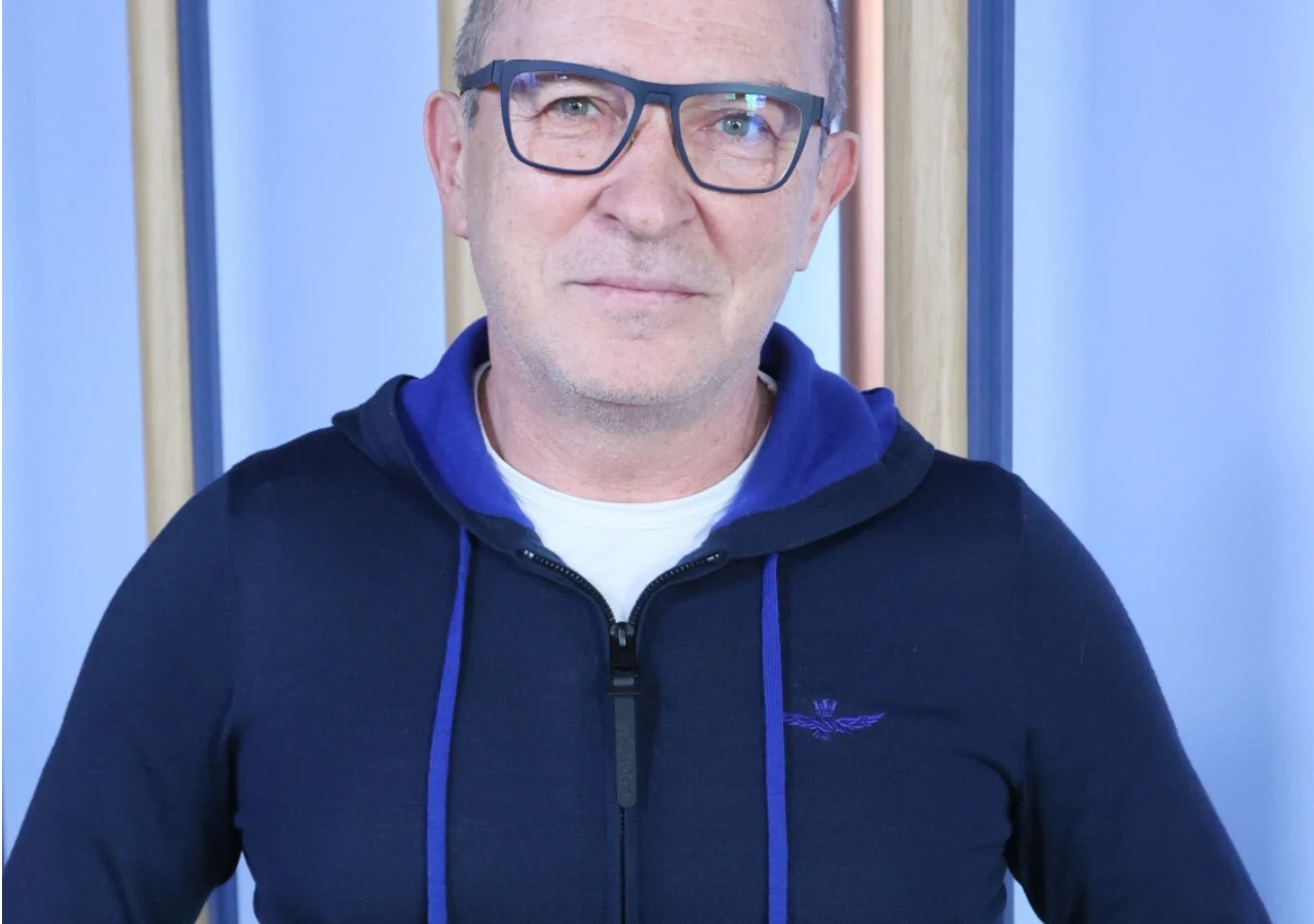 Andrzej Krzywy Mężczyzna w okularach i granatowej, rozpinanej bluzie z kapturem stoi na tle jasnoniebieskiej ściany z pionowymi, kolorowymi panelami.