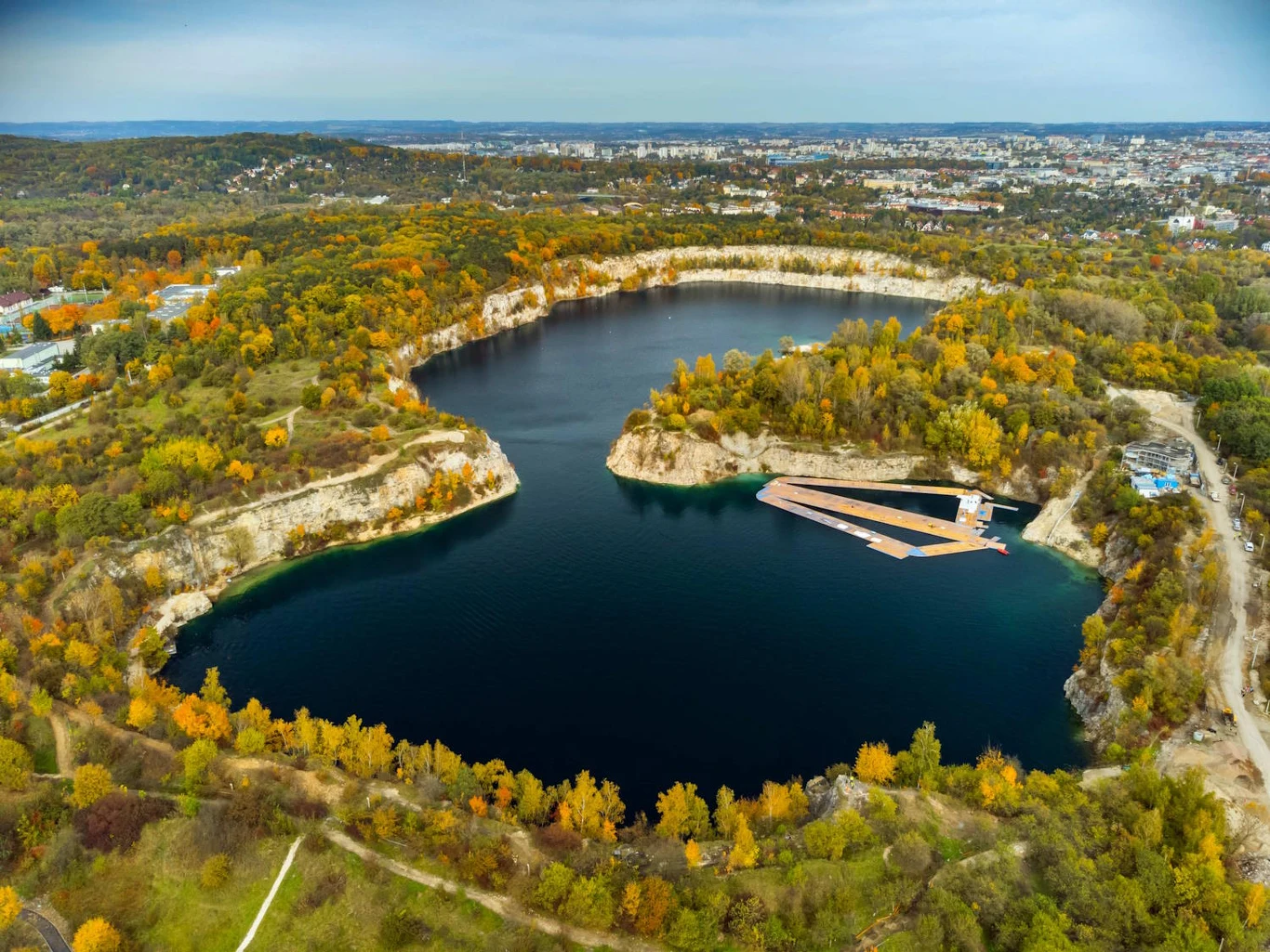 Zbiornik wodny otoczony stromymi, skalistymi brzegami porośniętymi kolorowymi jesiennymi drzewami, z widocznym nowoczesnym pomostem na jednym z brzegów oraz panoramą miasta w oddali.