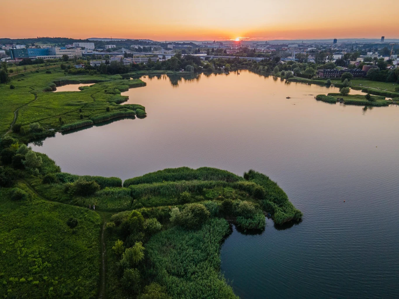 Jezioro otoczone zielenią, z widocznym zachodem słońca nad zabudowaniami miejskimi w tle.