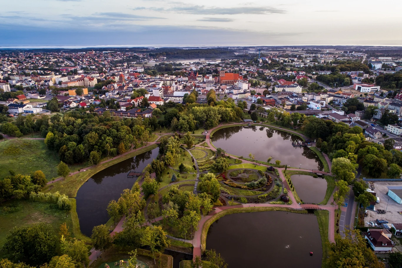 Park z dużymi stawami otoczonymi zielenią, ścieżki spacerowe, w tle zabudowa miejska z charakterystycznym czerwonym kościołem pośrodku panoramy miasta.