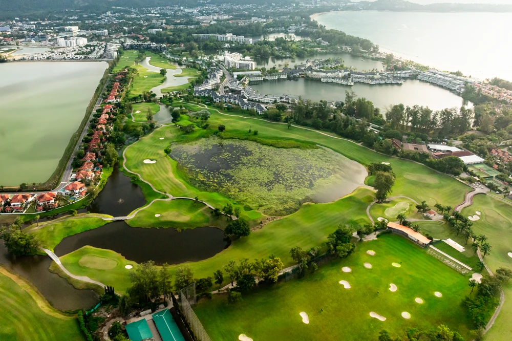 Kompleksowo zagospodarowane pole golfowe z licznymi zielonymi fairwayami, kilkoma dużymi stawami wodnymi oraz zabudowaniami mieszkalnymi i hotelowymi w tle, otoczone bujną roślinnością i pagórkowatym terenem.