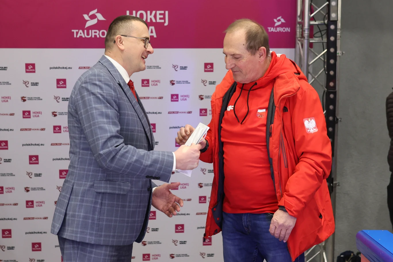 Dwóch mężczyzn podczas oficjalnego spotkania, jeden w garniturze wręcza dokument lub nagrodę drugiemu ubranemu w czerwoną kurtkę z emblematem Polski na piersi; w tle ścianka z logotypami sponsorów wydarzenia sportowego.