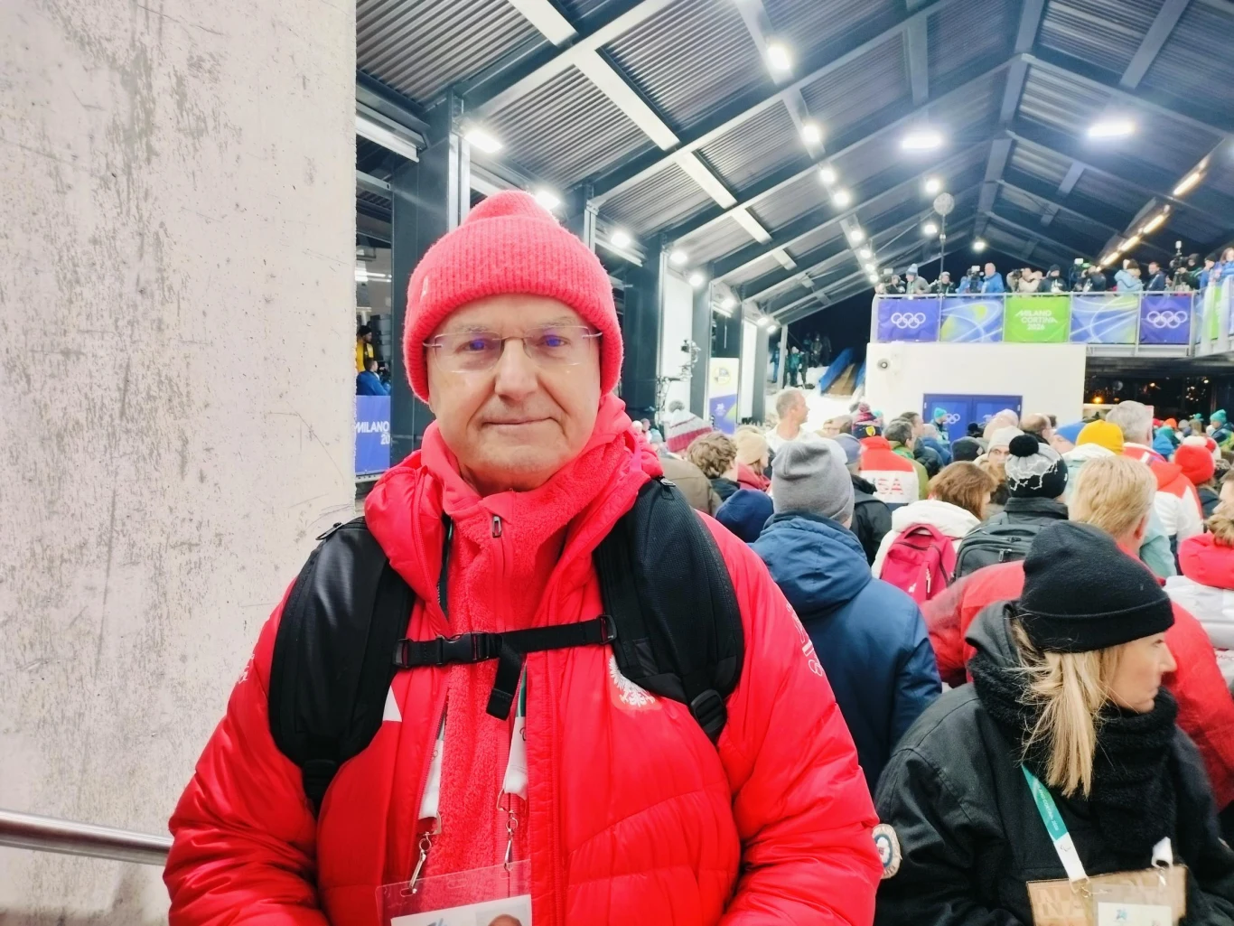 Mężczyzna w czerwonej kurtce i czapce na tle tłumu osób ubranych zimowo podczas wydarzenia sportowego w zadaszonej hali z widocznym logo igrzysk olimpijskich.