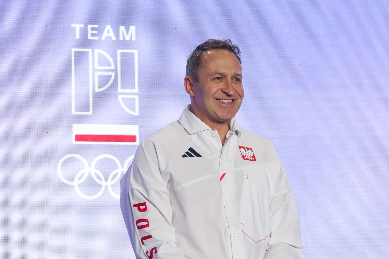 Mężczyzna w oficjalnej biało-czerwonej koszuli z logo Polski i napisem 'POLSKA' na tle grafiki z napisem 'TEAM POLAND', flagą Polski oraz symbolami olimpijskimi.