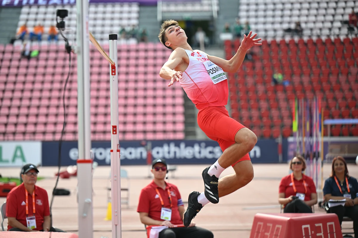 Lekkoatleta w czerwono-białym stroju wyskakuje w górę podczas konkurencji skoku wzwyż na stadionie lekkoatletycznym, obserwowany przez sędziów i oficjalnych pracowników zawodów.
