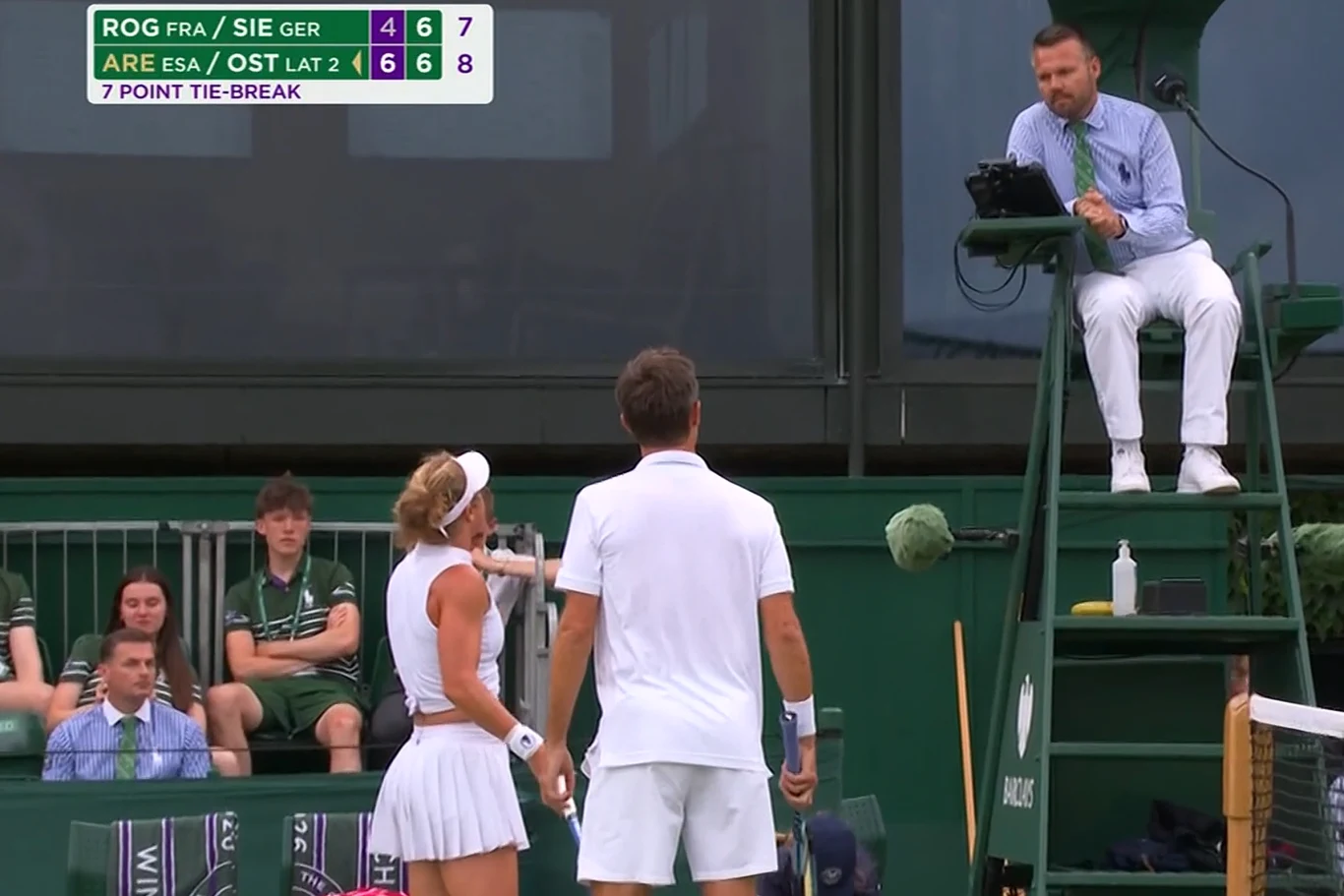 Laura Siegemund i Edouard Roger-Vasselin ruszyli z pretensjami do sędziego Para tenisistów w białych strojach prowadzi rozmowę z sędzią siedzącym na podwyższeniu podczas meczu na trawiastym korcie, w tle widoczni są sędziowie liniowi i widzowie, a na tablicy wyników wyświetlane są aktualne rezultaty spotkania.