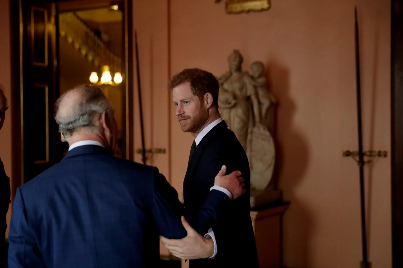 Dwóch mężczyzn ubranych w eleganckie garnitury, stojących w klasycznie urządzonym wnętrzu, w tle widoczna rzeźba oraz wysokie włócznie opierające się o ścianę.