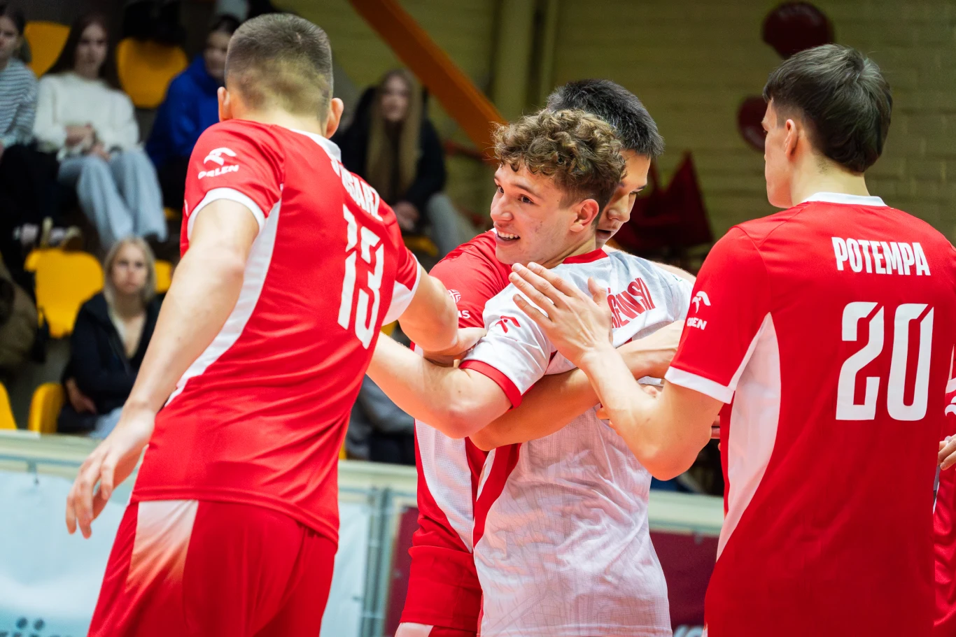 Grupa siatkarzy ubranych w czerwono-białe stroje świętuje zdobyty punkt, obejmując się i uśmiechając na boisku sportowym.