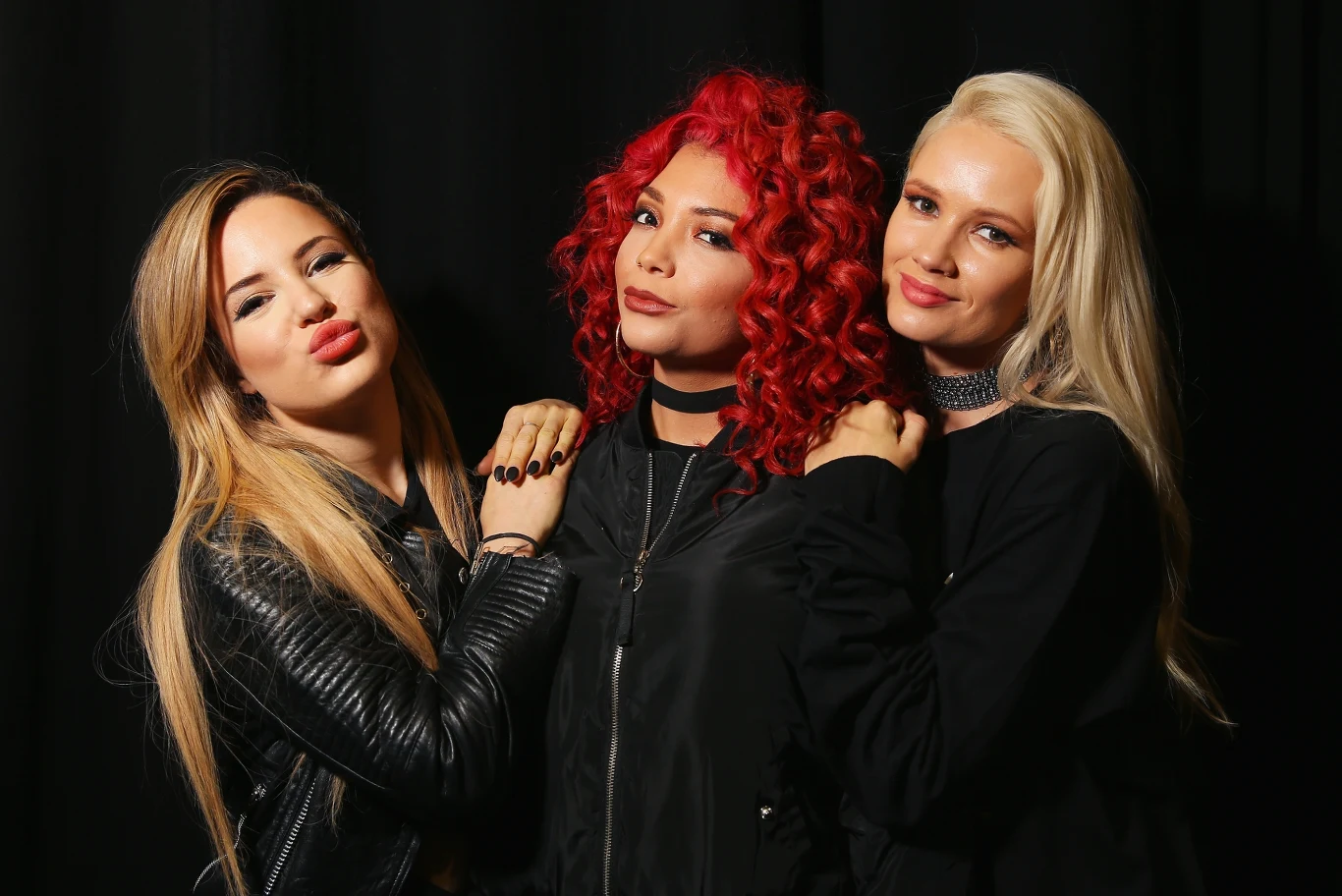 Natasha Slayton, Jazzy Mejia and Lauren Bennett z zespołu G.R.L Trzy młode kobiety w czarnych stylizacjach stoją obok siebie na tle ciemnej kurtyny, środkowa ma kręcone, czerwone włosy, po bokach blondynka i dziewczyna o jasnobrązowych włosach, wszystkie pozują uśmiechnięte i pewne siebie.