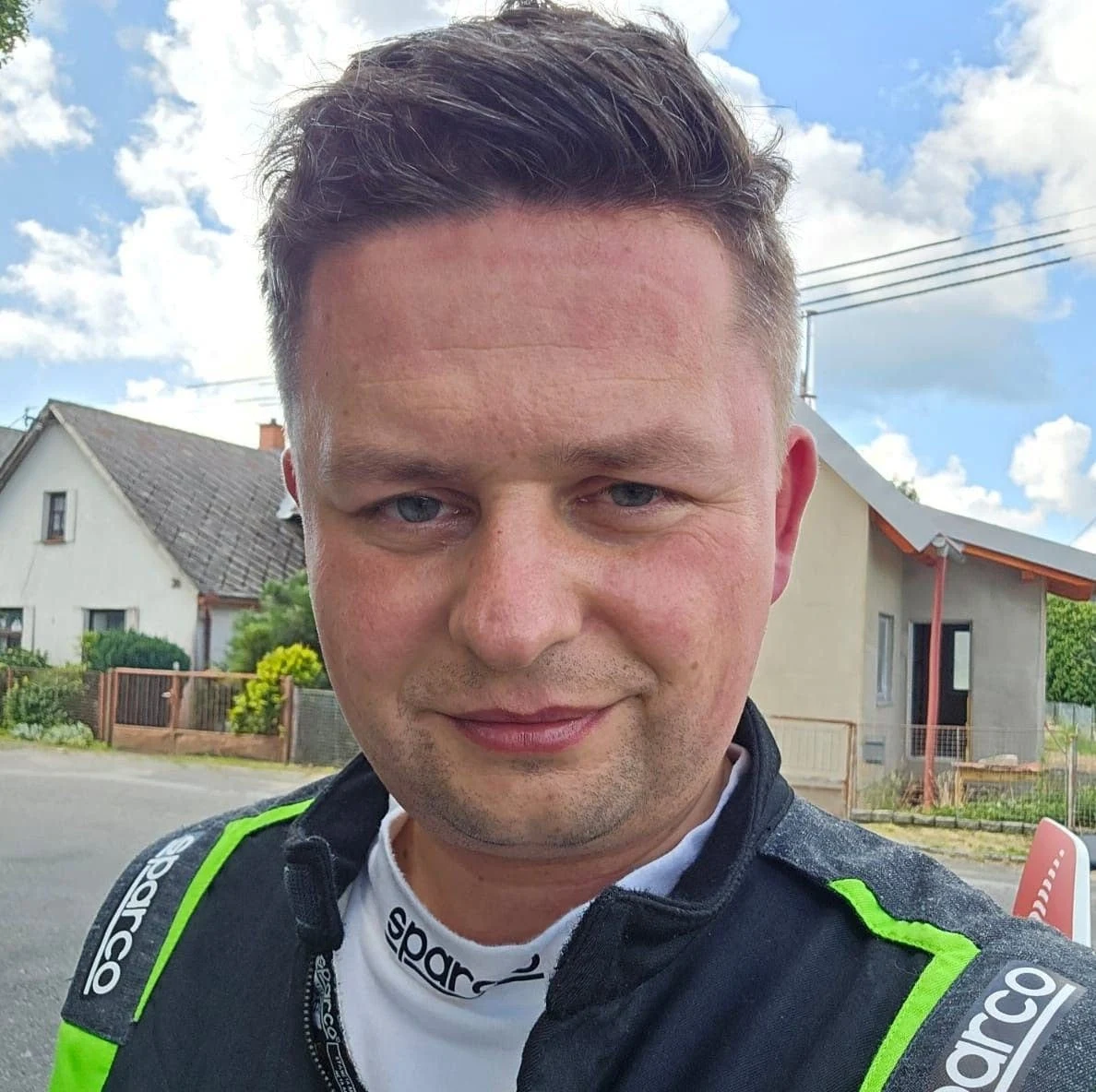 Mężczyzna ubrany w czarno-zielony kombinezon sportowy marki Sparco stoi na zewnątrz przed domami jednorodzinnymi, w tle widoczne niebo z białymi chmurami.